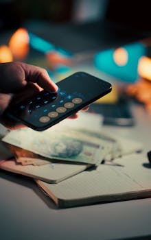 A hand holds a smartphone running a calculator app, with cash and a notebook on a desk, symbolizing budgeting and finance management.