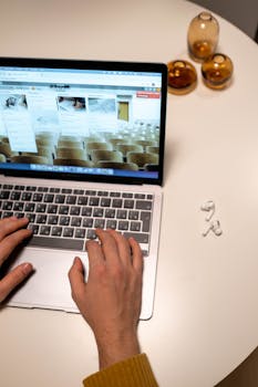 A person typing on a laptop at a white table with AirPods and glasses.