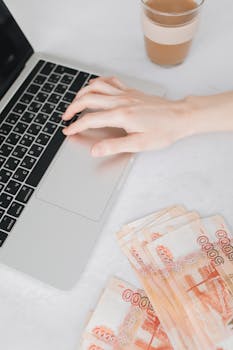 A person typing on a laptop next to a stack of Russian Rubles on a white table.