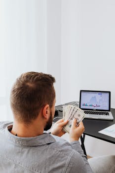 Businessman holding money while analyzing stock market data on laptop at home office setup.