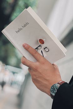 Close-up of a hand holding 'hello, habits' book with tea and watch image.