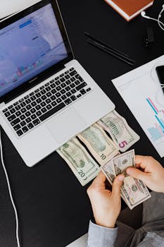 Person counting cash with laptop on desk, financial graphs visible.